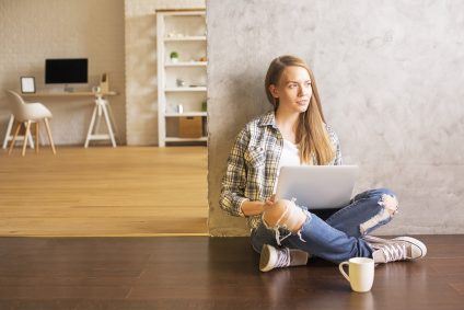 Young female sitting on wooden floor with coffee in interior and using laptop